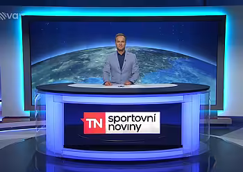 Moderátor stojí za moderním studiovým pultem s obrazovkou Země v pozadí.