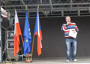 Moderátor v džínách a pruhovaném tričku stojí na pódiu s českými vlajkami v pozadí.