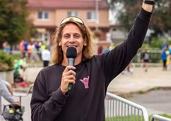 Moderátor v černé bundě drží mikrofon a fotoaparát venku při akci.