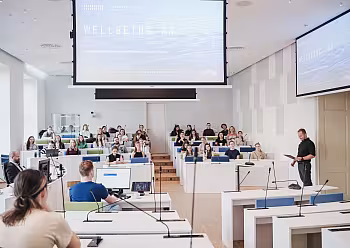 Moderátorka v tmavém oblečení stojí u pódia před publikem v moderní konferenční místnosti.