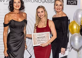 Tři ženy v elegantních šatech pózují s certifikátem před reklamní stěnou.