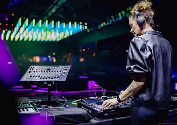 Moderátorka v tmavém oblečení za DJ pultem s barevným osvětlením v pozadí.