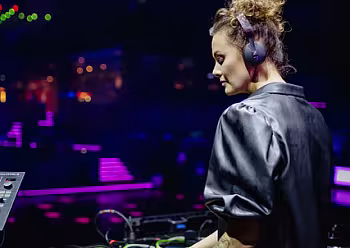 DJ v šedém tričku u mixážního pultu za fialového osvětlení.
