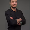 Moderátor v černém rolováku se založenýma rukama na šedém pozadí.