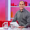 Moderátor v šedé košili sedí u stolu ve studiu s růžovým a červeným pozadím.
